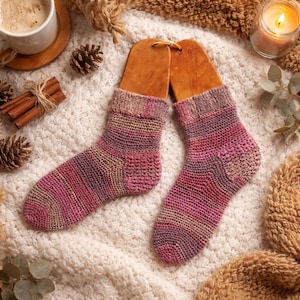 May include: A pair of hand-knitted socks in shades of pink, purple, and tan, displayed on wooden sock forms. The socks have a ribbed cuff and a striped pattern. The background includes a white knitted blanket, a cup of coffee, and a lit candle.