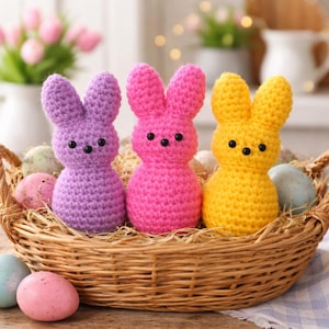 May include: Three handmade crochet bunny figures in purple, pink, and yellow sit in a woven basket filled with straw and speckled Easter eggs. The bunnies have black bead eyes and upright ears, perfect for Easter decor.