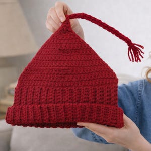 May include: A handmade, red crochet hat with a pointed top and a long, braided tassel. The hat features a ribbed brim and a textured stitch pattern. The hat is held up by a person.