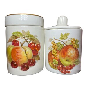 Puede incluir: Dos botes de cerámica blanca con tapas. Cada bote está decorado con un diseño de frutas de colores con cerezas, manzanas y otras frutas. Los botes tienen un borde dorado.