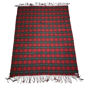 Puede incluir: Una manta a cuadros rojos y verdes con flecos. La manta presenta un patrón de tartán clásico con una cuadrícula repetida de líneas rojas y verdes. Los flecos son de color marrón oscuro.