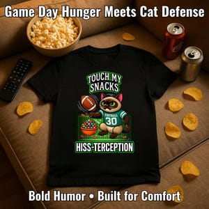 Peut inclure: T-shirt noir avec un chat de dessin animé portant un maillot de football, tenant un ballon et un bol de collations. Le texte sur le t-shirt dit "Touch My Snacks" et "Hiss-terception". L'image comprend des collations et des boissons.
