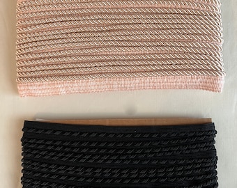May include: Two rolls of decorative trim. The top roll is a light pink and cream twisted cord trim. The bottom roll is black twisted cord trim. Both rolls are neatly stacked, showing the texture and pattern of the trim.