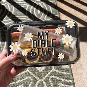 May include: A rectangular clear plastic zippered pouch with black trim, decorated with white and yellow daisy decals. The front features the black text MY BIBLE STUFF. Inside, various stationery items, including pens, small rolls of tape, and a beaded charm.
