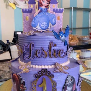 May include: A two-tiered purple cake decorated for a birthday. The top tier has the name "Leslie" in gold script. The cake is adorned with a princess, a castle, butterflies, a blue bird, a squirrel, a rabbit, and a number one. The cake sits on a gold cake board.
