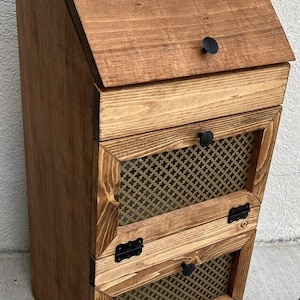 May include: A wooden storage cabinet with a slanted top and two mesh-fronted drawers. The cabinet is stained a warm brown color and features black hardware, including hinges and drawer pulls. The design is rustic and functional.