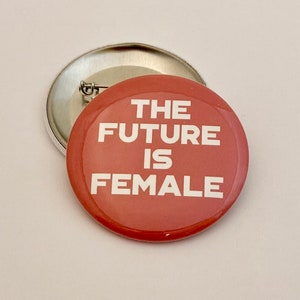 May include: A red button with the text "THE FUTURE IS FEMALE" in white. The button is round and has a pin on the back. Another button is partially visible in the background.