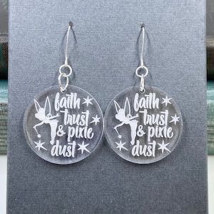 May include: A pair of clear acrylic earrings with silver-colored hooks. Each earring is a circle with the words "faith, trust & pixie dust" and a silhouette of a fairy. The text and fairy are white.