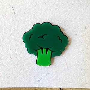 May include: A glossy green broccoli-shaped pin with a black border. The pin is set against a textured white background. The broccoli design features detailed florets and a stem. The pin is a fun, food-themed accessory.