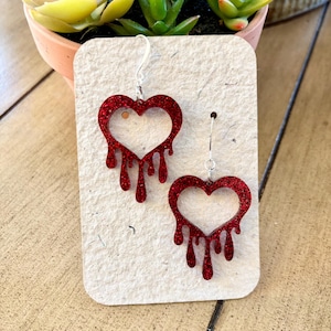 May include: A pair of red glitter heart earrings with a dripping design. The earrings are hanging on silver hooks and are displayed on a light brown card.