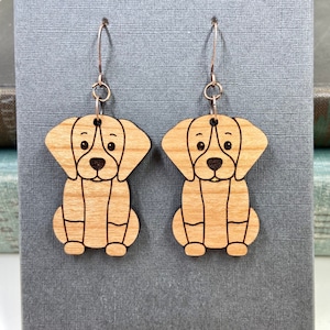 Puede incluir: Un par de pendientes de madera con forma de perro. Cada pendiente presenta un diseño de perro marrón claro cortado con láser con detalles en negro. Los pendientes cuelgan de ganchos de color plateado.