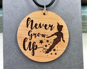 以下が含まれることがあります： 「Never Grow Up」の文字とピーターパンのシルエットが刻まれた、丸い木製ペンダントネックレス。ペンダントは明るい茶色で、濃い茶色のエッチングが施され、黒いコードで吊り下げられています。