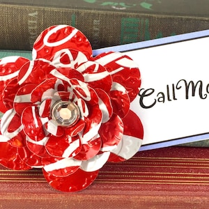 May include: A handmade red and white flower made from recycled materials, with a sparkling gem center. The flower is next to a white card with the words "Call Mom" in black script. The background includes vintage books.
