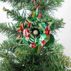 May include: A handmade Christmas ornament shaped like a flower, crafted from recycled aluminum cans. The ornament features red, green, and white colors, with a pearl-like bead in the center. It hangs from a red ribbon on a Christmas tree.