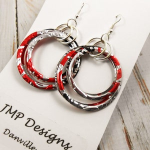May include: A pair of handmade hoop earrings. The earrings feature three interlocking circles made from recycled aluminum, with red, silver, and black patterns. The earrings hang from silver-colored ear wires. The text "JMP Designs Danville" is visible.