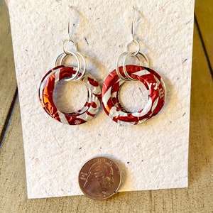 May include: A pair of handmade earrings featuring circular red and white patterned elements, connected by silver-toned hoops and hooks. The earrings are displayed on a textured, off-white surface, with a quarter coin for size comparison.