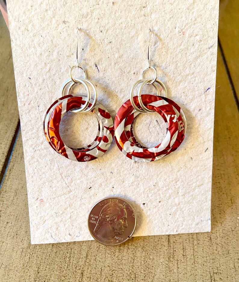 May include: A pair of handmade earrings featuring circular red and white patterned elements, connected by silver-toned hoops and hooks. The earrings are displayed on a textured, off-white surface, with a quarter coin for size comparison.