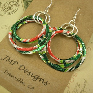 May include: A pair of handmade hoop earrings with a festive color scheme. The earrings feature three nested hoops in green, red, and white, with silver-tone ear wires. The earrings are made from recycled materials. The earrings are displayed on a brown surface.