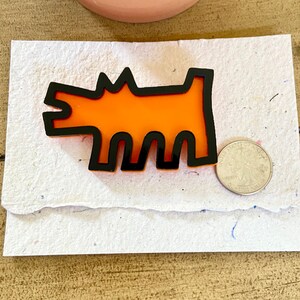 May include: Orange and black dog-shaped brooch. The brooch features a stylized dog design with an orange center and a black outline. A quarter coin is next to the brooch for size comparison. The brooch is on a white paper.