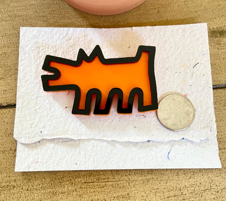 May include: Orange and black dog-shaped brooch. The brooch features a stylized dog design with an orange center and a black outline. A quarter coin is next to the brooch for size comparison. The brooch is on a white paper.