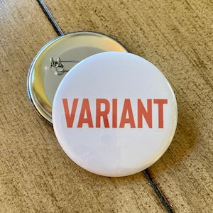May include: Two white round buttons with the word "VARIANT" in orange letters. One button is in front of the other, showing the pin back. The buttons are on a wooden surface.
