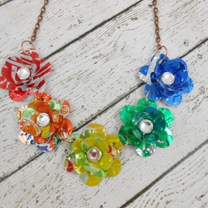 May include: A handmade necklace featuring five colorful flower pendants. Each flower is crafted from recycled materials, with a sparkling rhinestone center. The flowers are in shades of red, orange, yellow, green, and blue, connected by a copper chain.