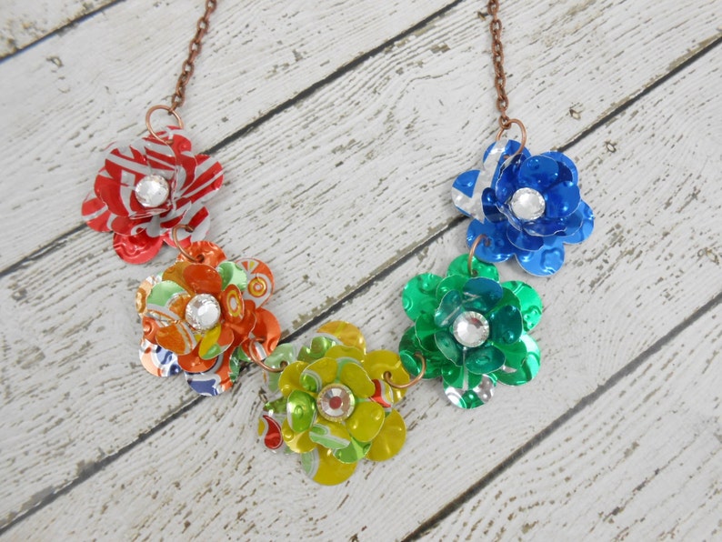 May include: A handmade necklace featuring five colorful flower pendants. Each flower is crafted from recycled materials, with a sparkling rhinestone center. The flowers are in shades of red, orange, yellow, green, and blue, connected by a copper chain.