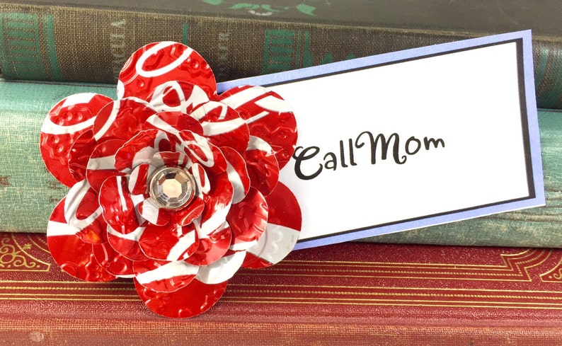 May include: A handmade red and white flower made from recycled materials, with a sparkling gem center. The flower is next to a white card with the words "Call Mom" in black script. The background includes vintage books.