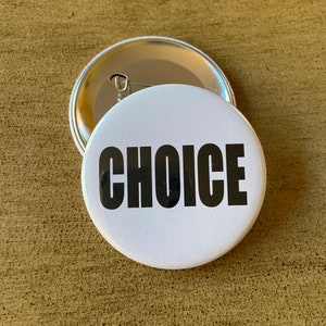 May include: Two white, circular badges with the word "CHOICE" in bold black text. One badge is on top of the other, revealing the pin back. The badges rest on a textured, light brown surface.