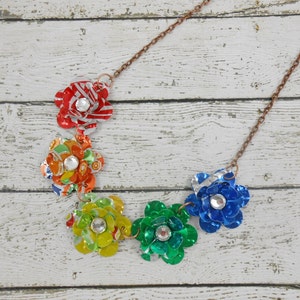 May include: A handmade necklace featuring five colorful flower pendants made from recycled materials. Each flower is a different color, including red, orange, yellow, green, and blue, and has a sparkling rhinestone center. The necklace has a copper-colored chain.