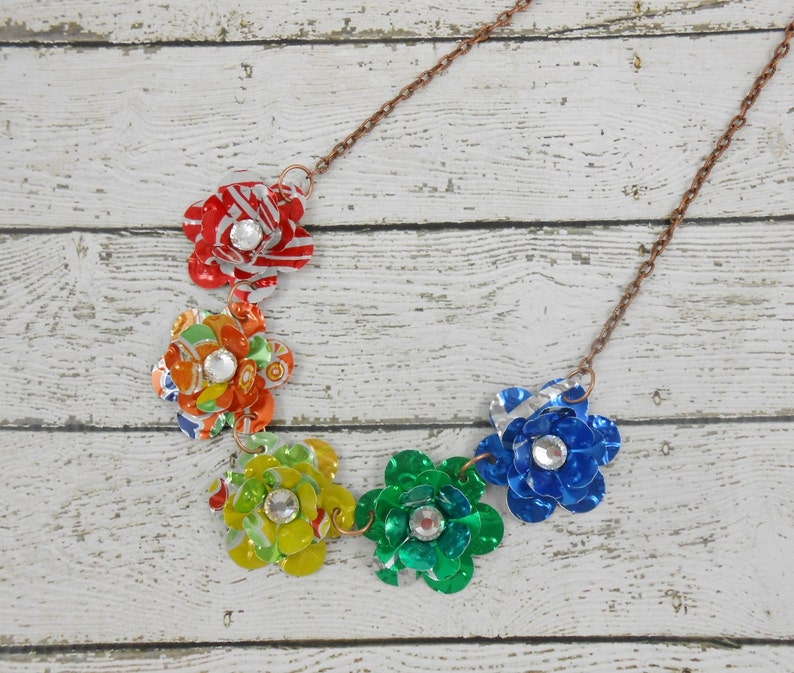 May include: A handmade necklace featuring five colorful flower pendants made from recycled materials. Each flower is a different color, including red, orange, yellow, green, and blue, and has a sparkling rhinestone center. The necklace has a copper-colored chain.