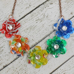 May include: A handmade necklace featuring five colorful flower pendants. Each flower is crafted from recycled metal and adorned with a sparkling rhinestone center. The flowers are in shades of red, blue, green, yellow, and orange, connected by a copper-colored chain.