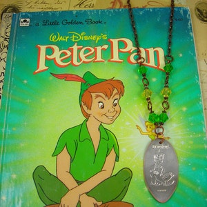 Puede incluir: Un libro de oro vintage "Peter Pan" se combina con un collar. El collar tiene un colgante plateado con un diseño de Peter Pan y un dije de Campanilla, cuentas verdes y amarillas y una cadena marrón. La portada del libro muestra a Peter Pan con ropa verde.