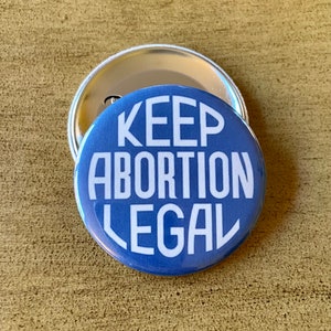 May include: A blue button with the text "KEEP ABORTION LEGAL" in white. The button is round and has a silver metal backing. The button is on a wooden surface.