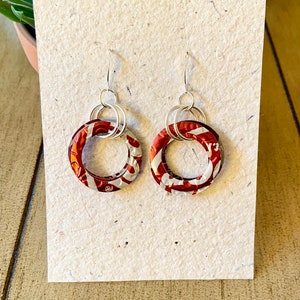 May include: A pair of handmade earrings featuring circular pendants made from recycled aluminum cans. The pendants have a red and white pattern, and are connected to silver-colored ear wires. The earrings are displayed on a textured, off-white card.