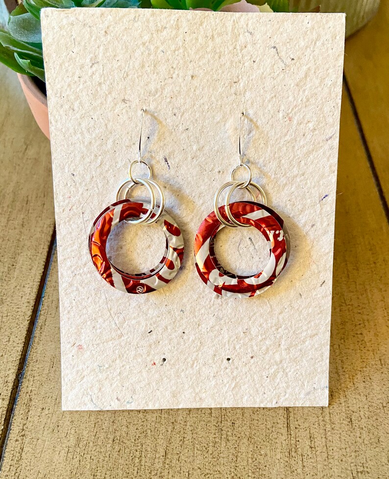 May include: A pair of handmade earrings featuring circular pendants made from recycled aluminum cans. The pendants have a red and white pattern, and are connected to silver-colored ear wires. The earrings are displayed on a textured, off-white card.