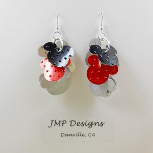 Puede incluir: Un par de pendientes colgantes con un dise&ntilde;o de Mickey Mouse. Cada pendiente tiene una cabeza de Mickey Mouse negra y roja, rodeada de acentos circulares plateados. Los pendientes est&aacute;n suspendidos de ganchos plateados. El texto "JMP Designs Danville, CA" es visible.