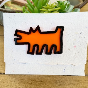 May include: An orange dog-shaped pin with a black outline is displayed on a textured, off-white card. The dog has a simple, stylized design. The card is layered and has a torn edge. The background includes a succulent plant and a wooden surface.