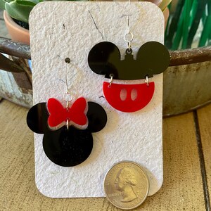 May include: A pair of black and red acrylic earrings featuring Mickey Mouse and Minnie Mouse designs. One earring is Mickey's face with a red bottom and white accents. The other earring is Minnie's face with a red bow. The earrings are displayed on a white card.