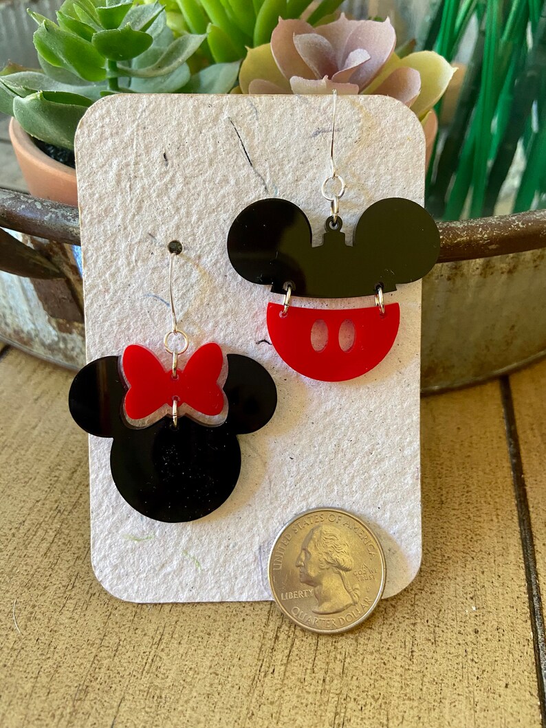 May include: A pair of black and red acrylic earrings featuring Mickey Mouse and Minnie Mouse designs. One earring is Mickey's face with a red bottom and white accents. The other earring is Minnie's face with a red bow. The earrings are displayed on a white card.
