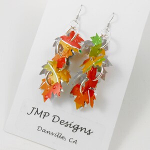 May include: Dangle earrings with a leaf design in autumnal colours. The leaves are in shades of red, orange, yellow, and green, with silver-tone metal accents. The earrings are displayed on a white card with the text "JMP Designs Danville, CA."
