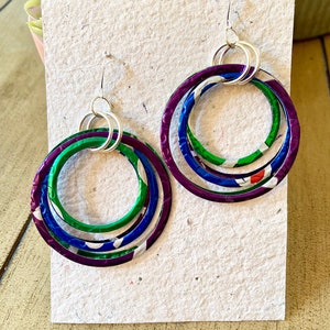May include: A pair of handmade hoop earrings with a layered design. The earrings feature three nested hoops in purple, green, and blue, with silver-tone ear wires. The hoops have a textured, metallic appearance, adding a unique touch to the jewelry.