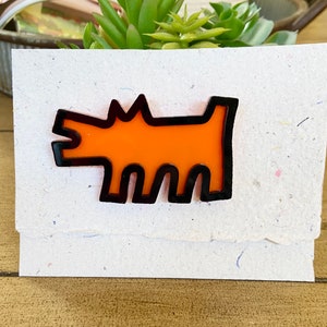 May include: An orange dog-shaped pin with a black outline is displayed on a textured, off-white card. The pin has a minimalist design, and the card has a torn edge. The background includes a small succulent plant and a wooden surface.