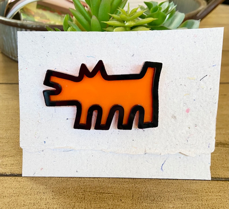 May include: An orange dog-shaped pin with a black outline is displayed on a textured, off-white card. The pin has a minimalist design, and the card has a torn edge. The background includes a small succulent plant and a wooden surface.