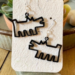 May include: A pair of black dog-shaped earrings. The earrings are cut-out designs with a simple outline of a dog. They hang from gold-colored ear wires. The earrings are displayed on a textured, off-white surface.