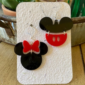 May include: A pair of black and red acrylic earrings. One earring features a black Mickey Mouse head with a red mouth. The other earring is a black Minnie Mouse head with a red bow. Both earrings have silver-coloured hooks.