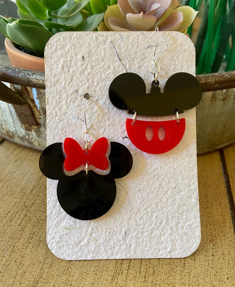 May include: A pair of black and red acrylic earrings. One earring features a black Mickey Mouse head with a red mouth. The other earring is a black Minnie Mouse head with a red bow. Both earrings have silver-coloured hooks.