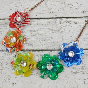 May include: A handcrafted necklace featuring six colorful flower pendants made from recycled aluminum cans. Each flower has a sparkling rhinestone center and is connected by a copper-colored chain. The flowers are red, orange, yellow, green, blue, and silver.