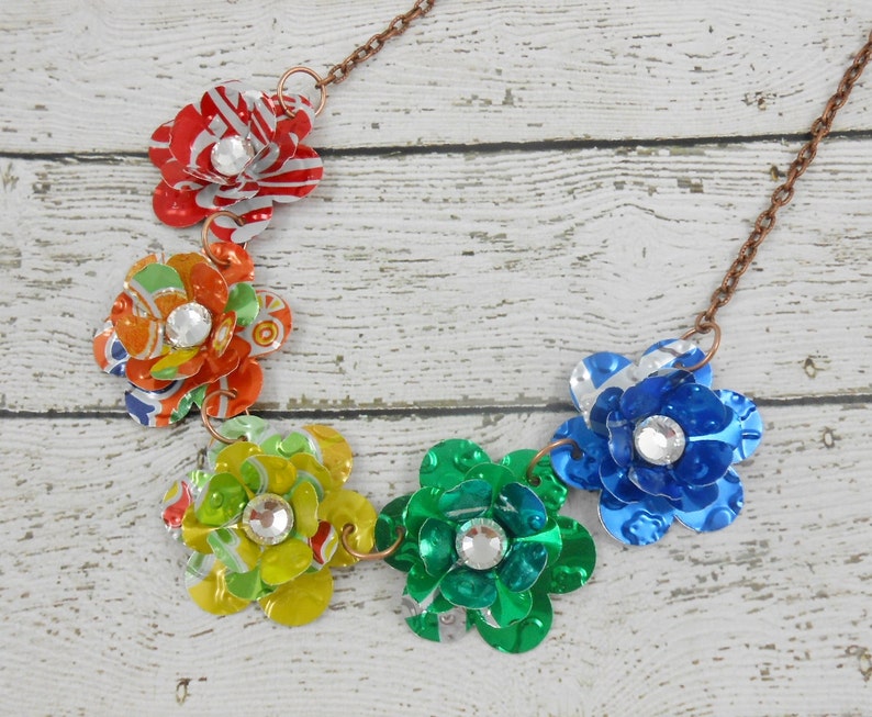 May include: A handcrafted necklace featuring six colorful flower pendants made from recycled aluminum cans. Each flower has a sparkling rhinestone center and is connected by a copper-colored chain. The flowers are red, orange, yellow, green, blue, and silver.