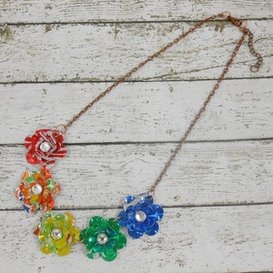 May include: A handmade necklace featuring six colorful flower pendants made from recycled materials. Each flower is a different color, including red, orange, yellow, green, and blue, and has a sparkling gem in the center. The chain is a copper color.
